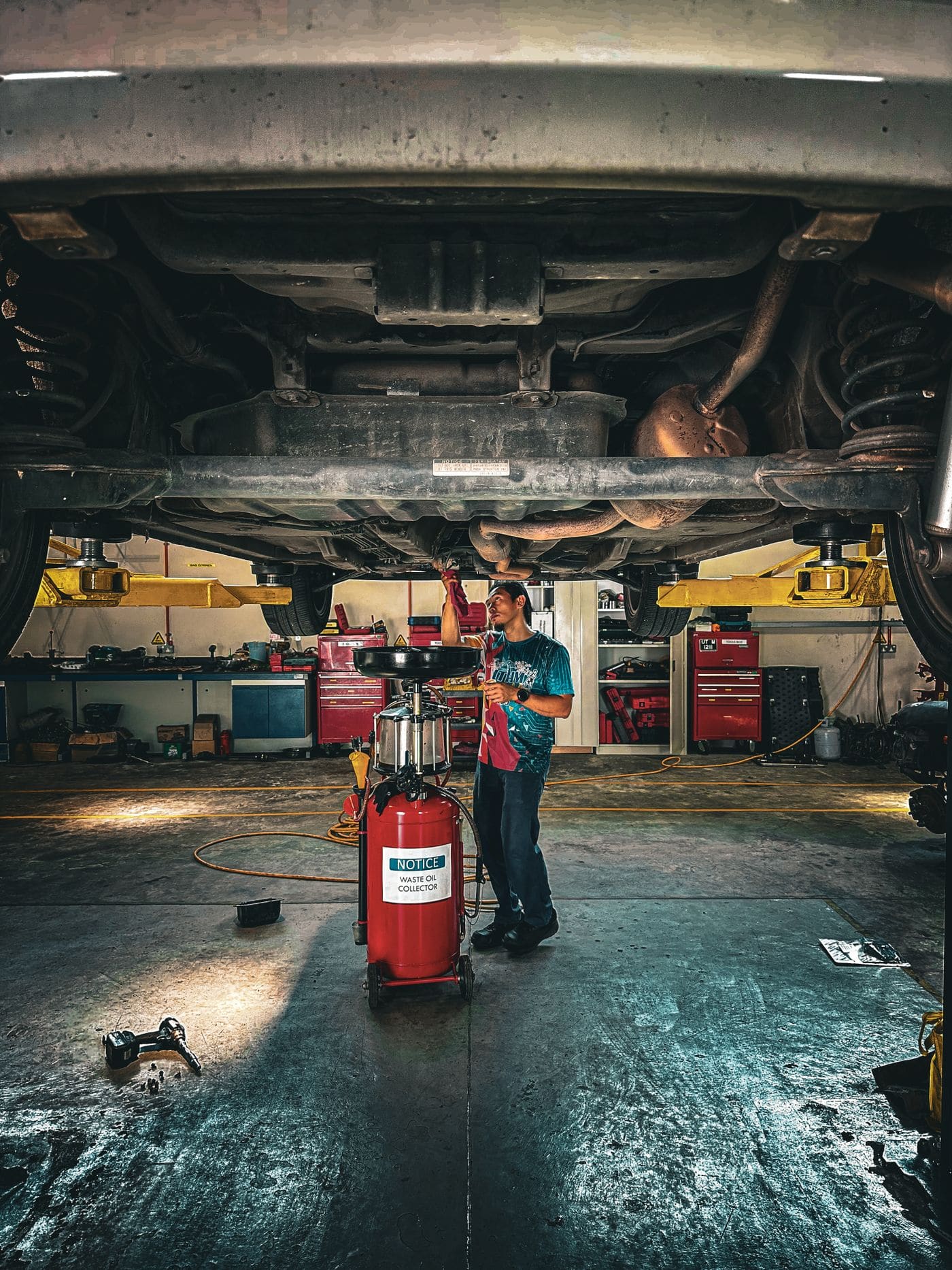 A mechanic working underneath a car raised on a two-post lift, surrounded by red tool chests in a repair bay
